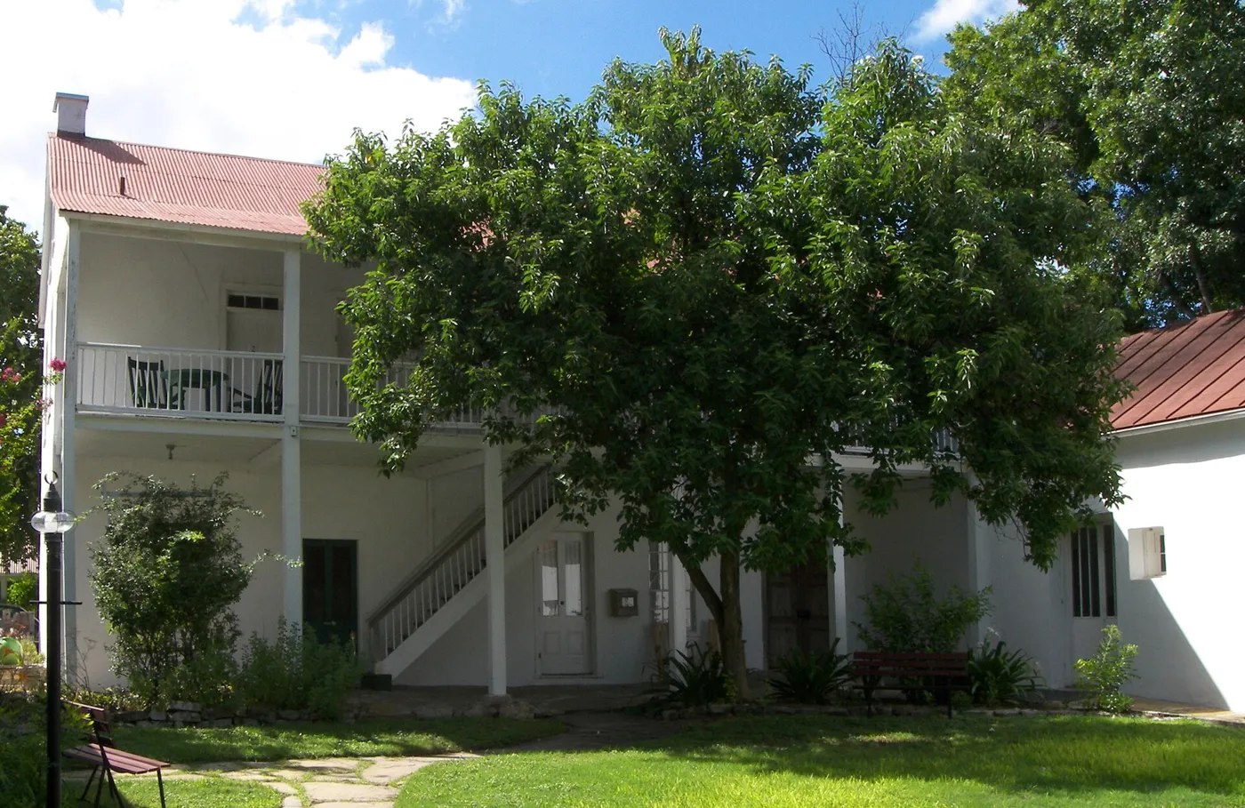 A white two-story stone inn with a long upper balcony and a red metal roof behind a shade tree